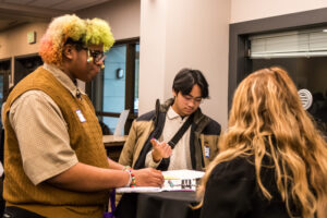 A man with a coat slung over his shoulders clicks a pen as two others face him.