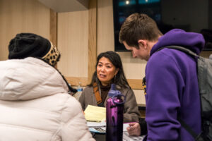 A woman with long dark hair speaks as two others listen.