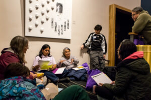 A group of people seated on a floor talk and look over pieces of paper.