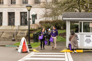 Several people with purple bags step into a crosswalk next to a small security kiosk.