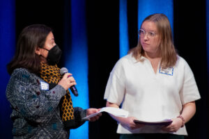 A woman in a black mask speaks into a handheld microphone while onother woman looks at her.