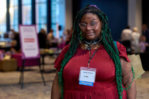 A woman with long teal dreadlocks, wearing glasses and a red dress, smiles at the camera.