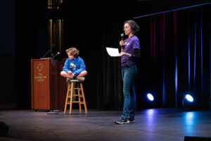 A woman on a stage speaks into a microphone held in one hand while clutching a sheet of paper as a small boy sits cross-legged on a stool nearby.
