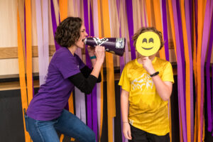 A woman in a purple shirt pretends to scream through a blowhorn directly at a small boy in a yellow T-shirt who's covering his face with a yellow smiley face prop.
