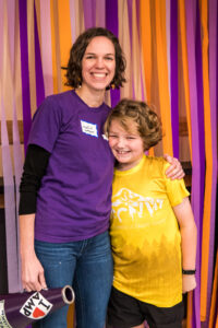 A woman in a purple shirt hugs a child in a yellow T-shirt with one arm while holding a purple blow horn in the other.