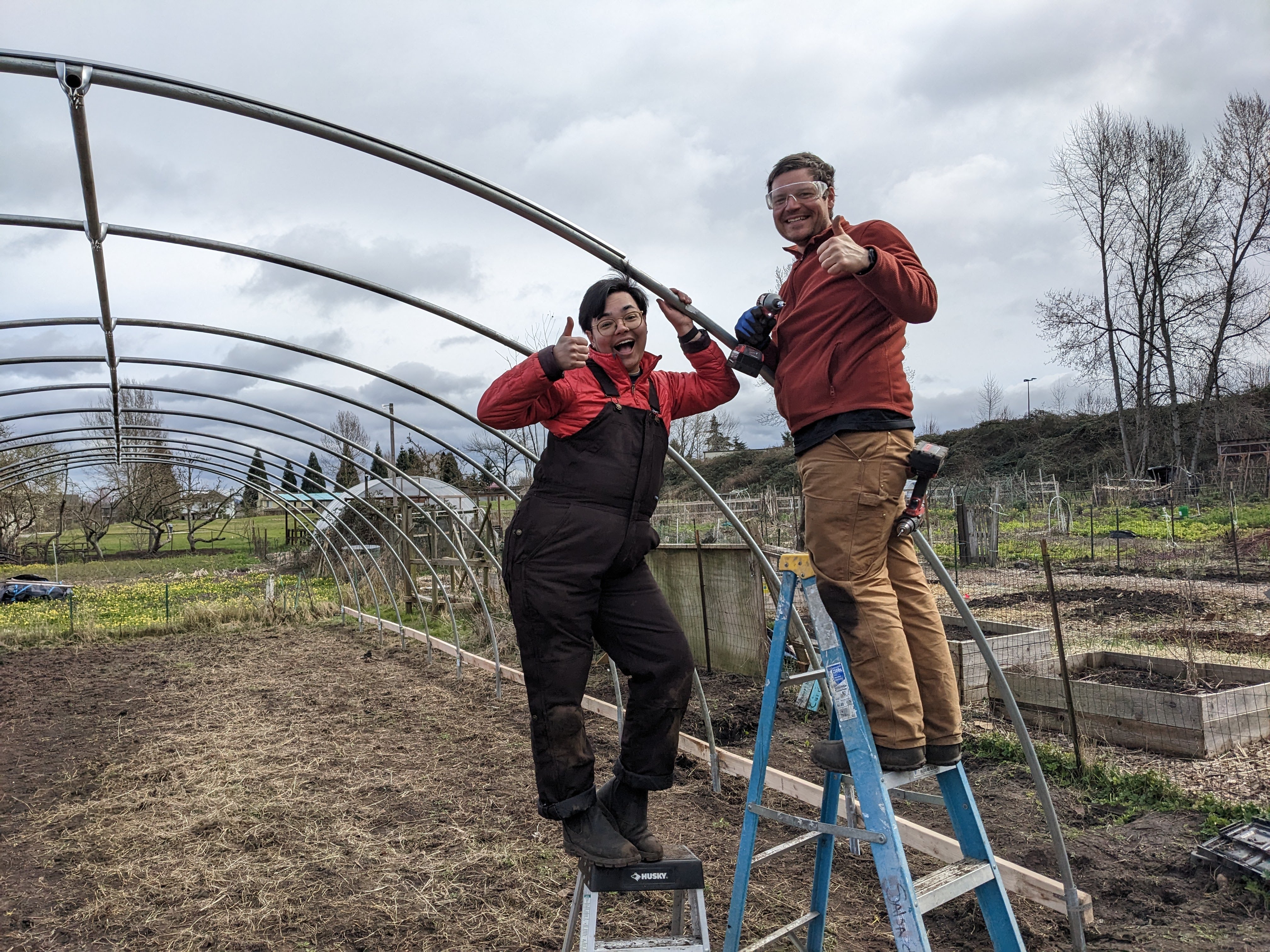two people on ladders give the thumbs up while working on a series of arched beams that will form the structure of a hoop house.
