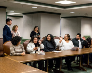 Several people seated along a wooden table raise their hands while looking up at a man standing off to the side.