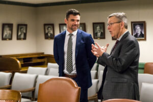 A man in a suit speaks as he gestures with his hands while another man in a suit and tie looks on.