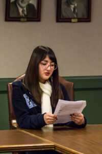 A woman in a scarf sits at the edge of a wooden table and carefully studies a sheet of paper in her hands.