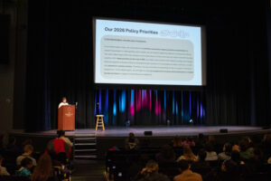 A speaker stands at a podium on a stage next to a large screen that reads "Our 2026 Policy Priorities" followed by a long paragraph of text.
