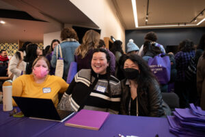 Three people sit at a long table in front of a group of other people.