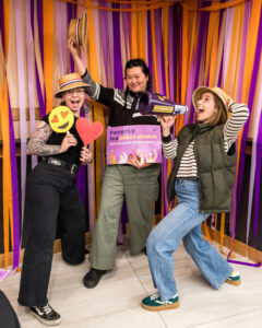 Three people strike goofy poses while holding props, including a sign that says "Poverty is a policy choice."