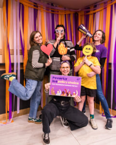 Five people strike goofy poses while holding props, including a sign that says "Poverty is a policy choice."