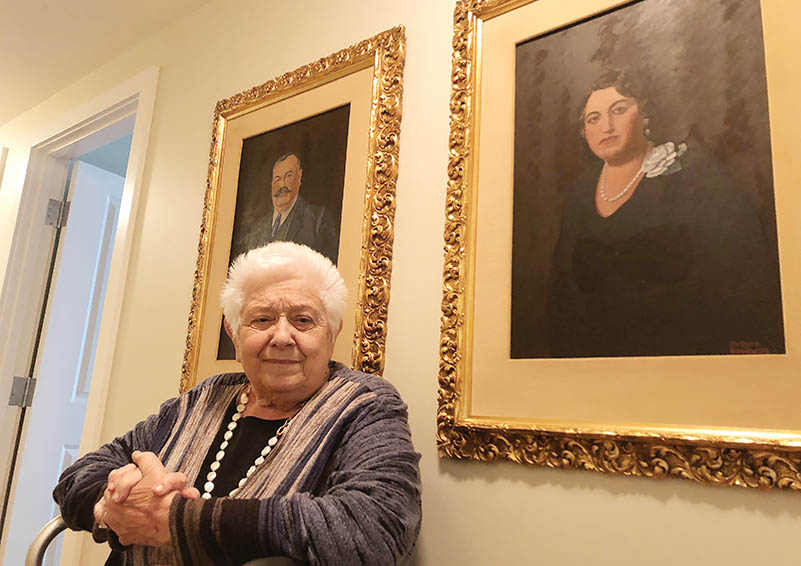 Elderly white woman seated in front of two large portraits of her parents