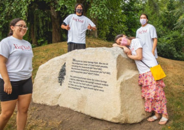 Several Indigenous youth standing around a rock carved with an inscription about the history and significance of Licton Springs.