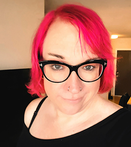 White woman with candy-red hair wearing black-rimmed glasses and a black top