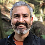Headshot of a smiling Latinx man, outdoors, with greying black hair and beard