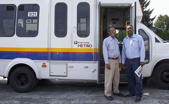 Two Black men in light blue work shirts smile in front of an accessible transportation bus