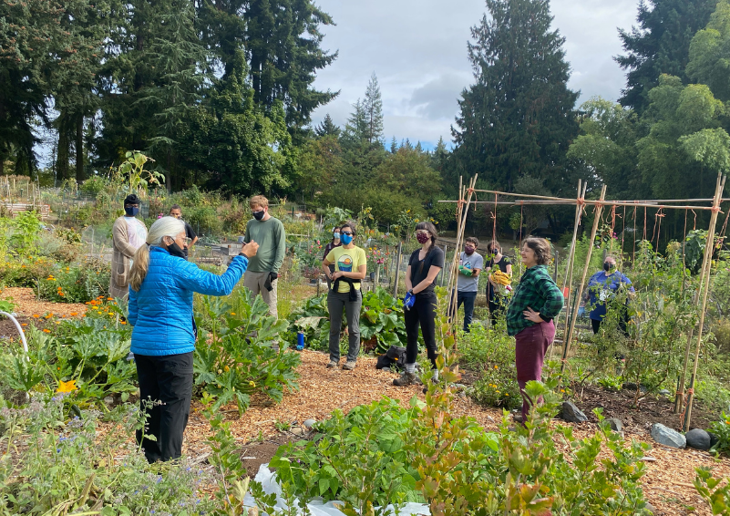 A woman in a blue coat speaks to group of people scattered around a large community garden in full bloom.