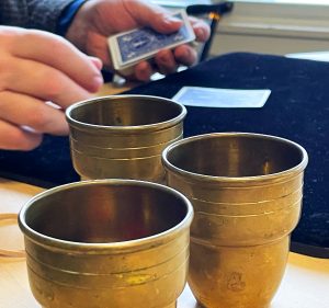 Three brass cups in the foreground with a pair of hands dealing cards behind them.