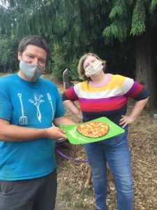 Two people standing outdoors wearing masks smiling, one is holding a homemade pizza