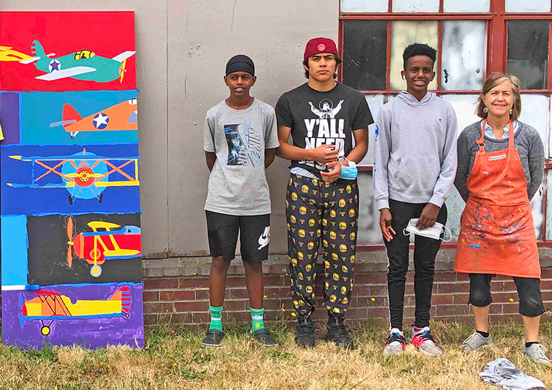 Three teenagers stand between a colorful mural and the artists who helped them create it.