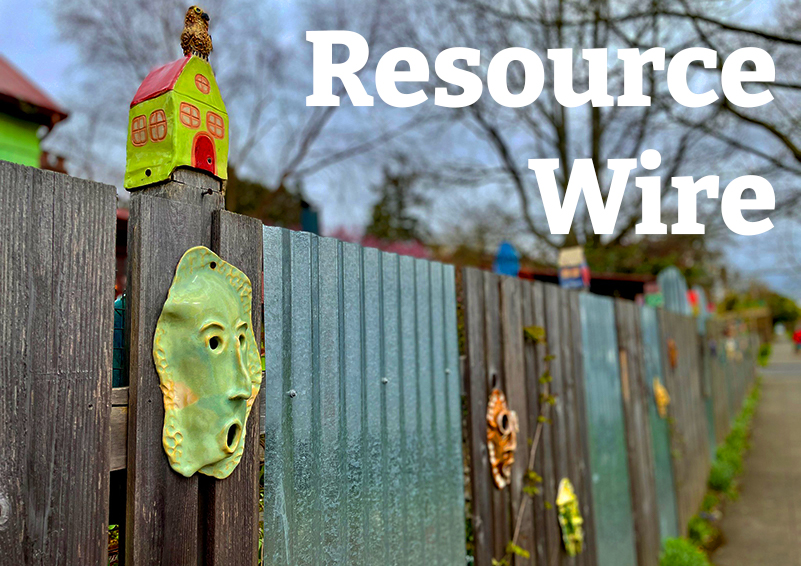 Colorfully painted clay faces and tiny homes posted on a wooden face facing a sidewalk.