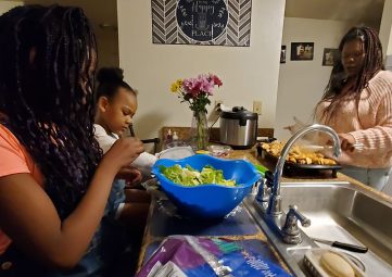 Two girls make salad in a bright blue bowl while their mom grills veggies and chicken