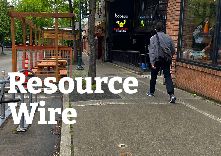 A man walking down a sidewalk lined with storefronts on one side and picnic tables in the street on the other. The words "Resource Wire" are embossed over the image.