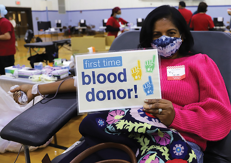 An Indian woman in a mask, neon pink top and floral skirt gives blood in a gymnasium. She holds a sign reading FIRST TIME blood donor!