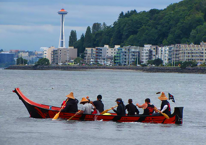Members of the Duwamish tribe paddle a traditional red-and-black canoe through the waters of Elliot Bay with condos and at Alki and the Space Needle in the background.