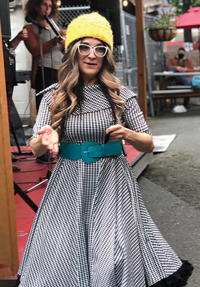 Woman dances down the street. She has long blondish hair and wears a yellow knitted cap, white-rimmed glasses, and a black & white checkered dress with an aqua belt.