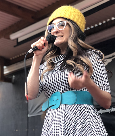 Woman on stage speaks into a mic. She has long blondish hair and wears a yellow knitted cap, white-rimmed glasses, and a black & white checkered dress with an aqua belt.