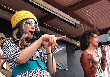 Two people perform on stage speaking into mics. The woman on the left has long blondish hair and wears a yellow knitted cap, white-rimmed glasses, and a black & white checkered dress with an aqua belt. The person on the right has curly brown hair and wears a tank top.