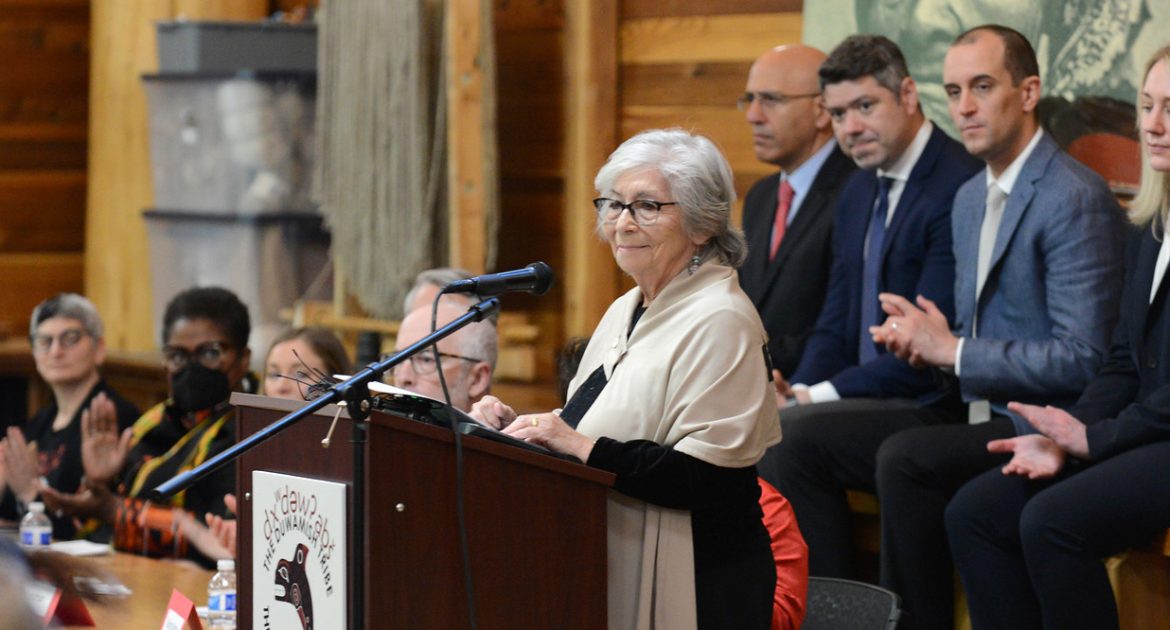 An older woman in white shall and white hair, stands at a lectern flanked by men in suits and others off behind.
