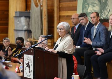 An older woman in white shall and white hair, stands at a lectern flanked by men in suits and others off behind.