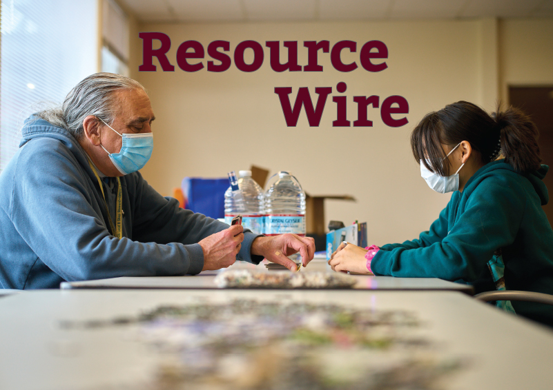 An older man and young girl play cards across a long table with the words "Resource Wire" above them.