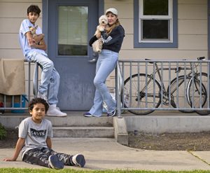 A teen and his mom holding dogs, leaning on the railing in front of a townhouse door. Bikes are parked on the porch. A younger boy sits in the grass in front of them.