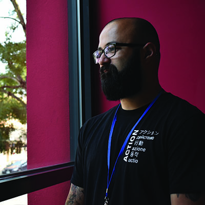 A man wearing dark-rimmed glasses, a black mustache and beard, and a black T-shirt with white lettering, stands in front of a red wall and gazes out a window.