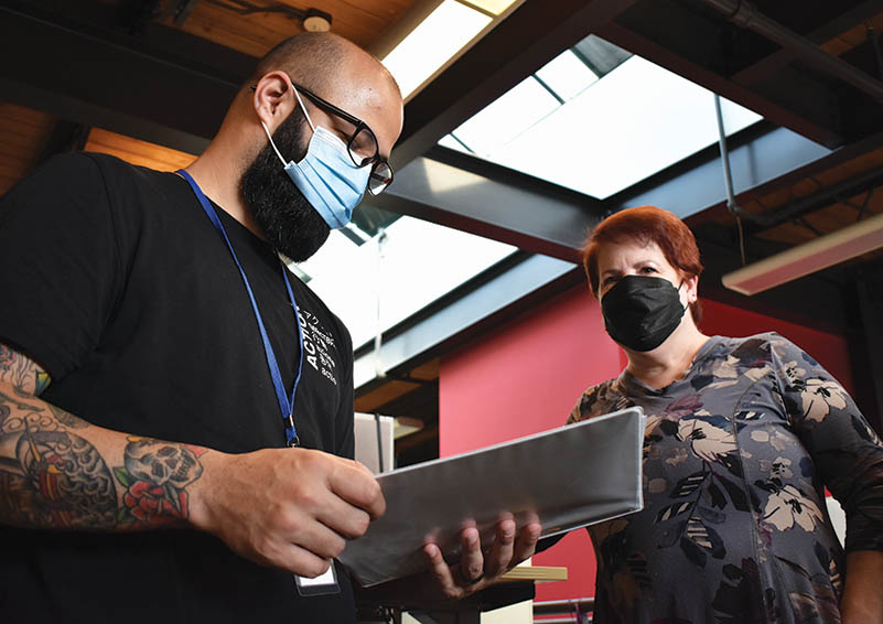 Two people under a skylight in an office with red walls. The man on the left looks at a folder. He wears a blue surgical mask, glasses, and a dark a beard, and has a forearm tattoo. The woman on the right has red hair and wears a floral shirt and a black mask.