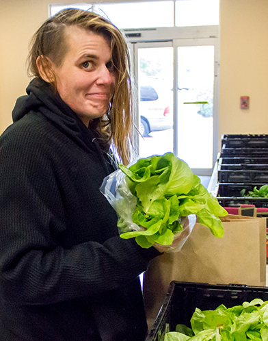 A person with blonde hair and a left side shave wearing all black smiles while holding a head of lettuce in their hand.