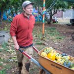 A man in a red hoodie and baseball hat pushes a wheelbarrow full of green and yellow squashes.