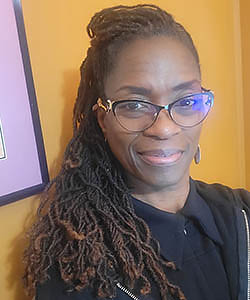 Headshot portrait of a Black woman with long braids and glasses in front of mango-colored wall.