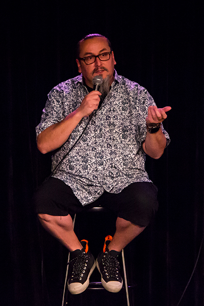 A man wearing black shorts and a patterned shirt sits on a stool and gestures while holding a microphone.