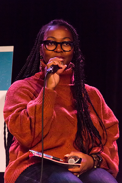 A woman with long braided hair and a loose red sweater talks into a microphone.