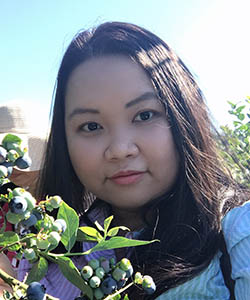 Headshot portrait of a young Asian woman with long black hair, posing with some blueberry bushes