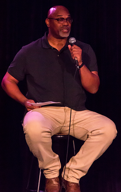 A man in in khakis and a dark shirt sits on a stool and speaks into a microphone.