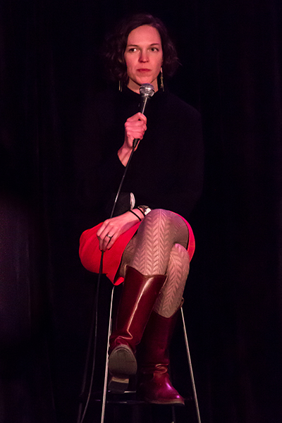A woman in a skirt and tall boots sits on a stool and speaks into a microphone.