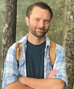 Portrait of a young white man with brown hair, mustache and beard, standing in a grove of trees. wearing a plaid shirt and backpack
