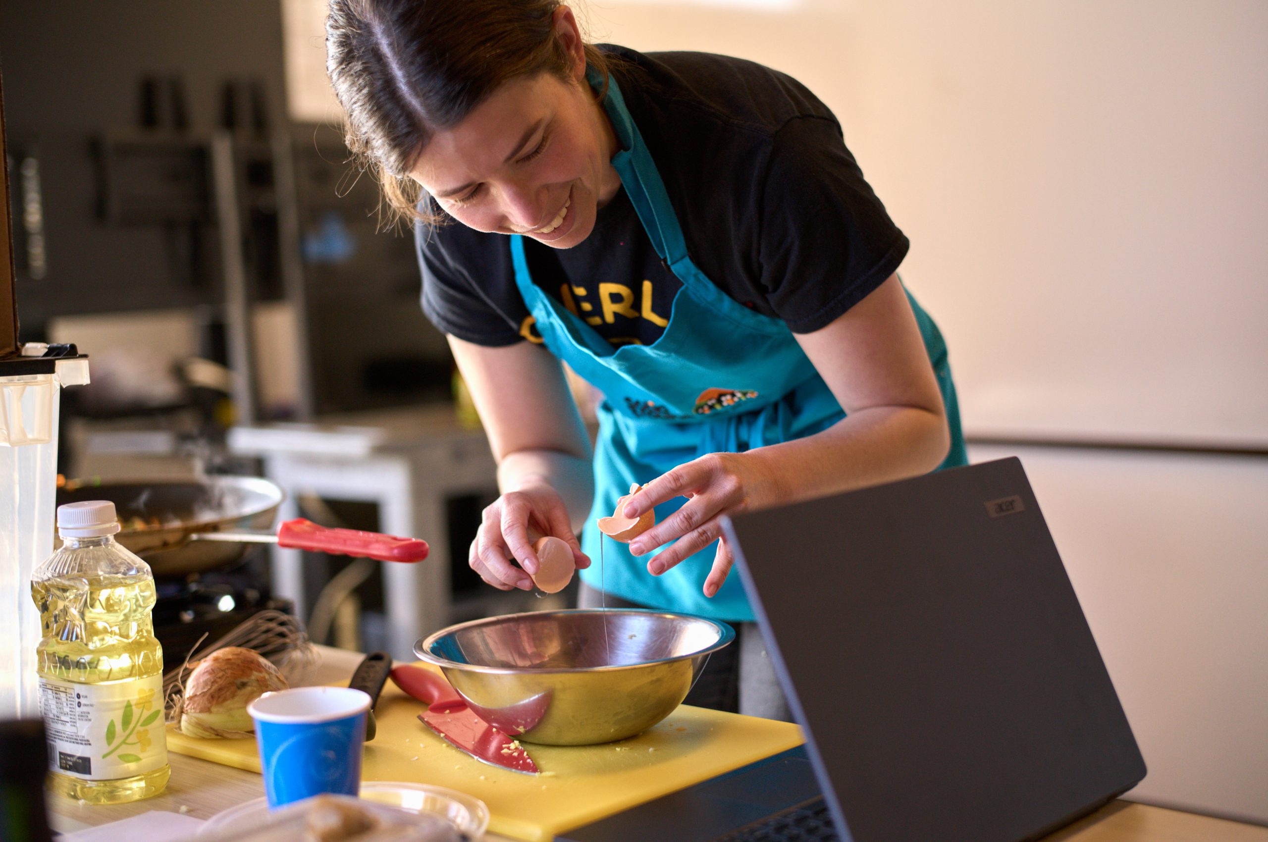 A woman with dark hair pulled back, wearing a dark T-shirt and aqua apron, cracks an egg into a bowl.
